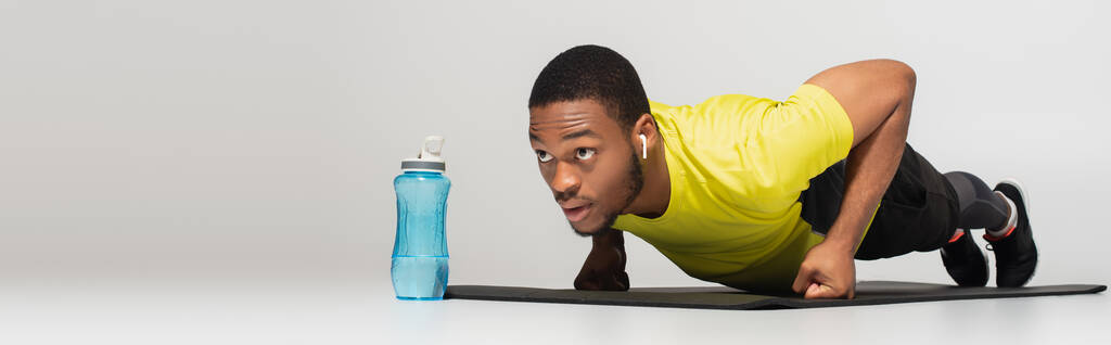 σπορ αφρικάνος Αμερικανός με ακουστικά κάνει πους απς στο στρώμα γυμναστικής κοντά σε αθλητικό μπουκάλι σε γκρι, banner - Φωτογραφία, εικόνα