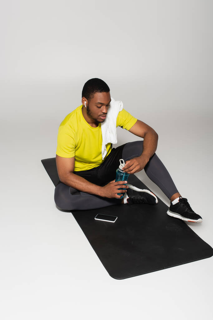 longitud completa del deportista afroamericano sentado en la colchoneta de fitness cerca de la botella de deportes y el teléfono inteligente en gris - Foto, Imagen