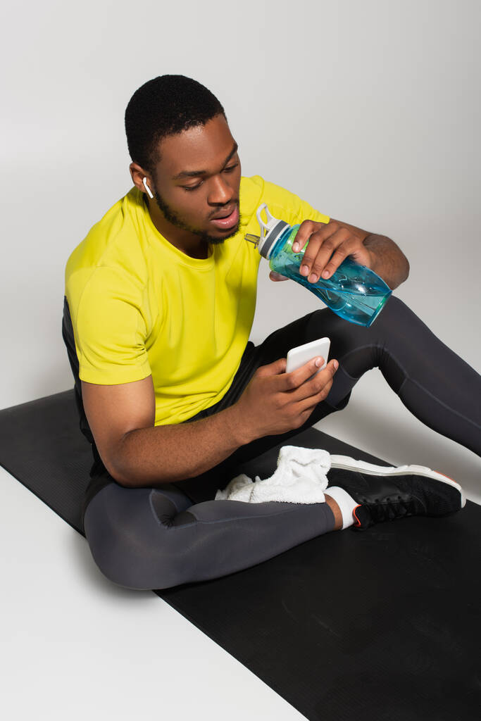 Αφροαμερικανός αθλητής που κάθεται σε στρώμα γυμναστικής και χρησιμοποιεί smartphone κοντά σε αθλητικό μπουκάλι σε γκρι  - Φωτογραφία, εικόνα