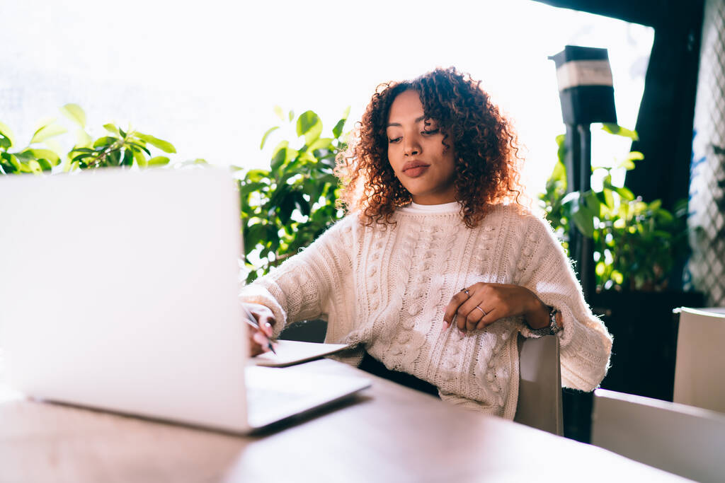 Bedachtzame Afro-Amerikaanse vrouw in casual kleding zitten aan houten tafel in een modern café en notities maken in het kopieerboek tijdens het werken op laptop op afstand - Foto, afbeelding