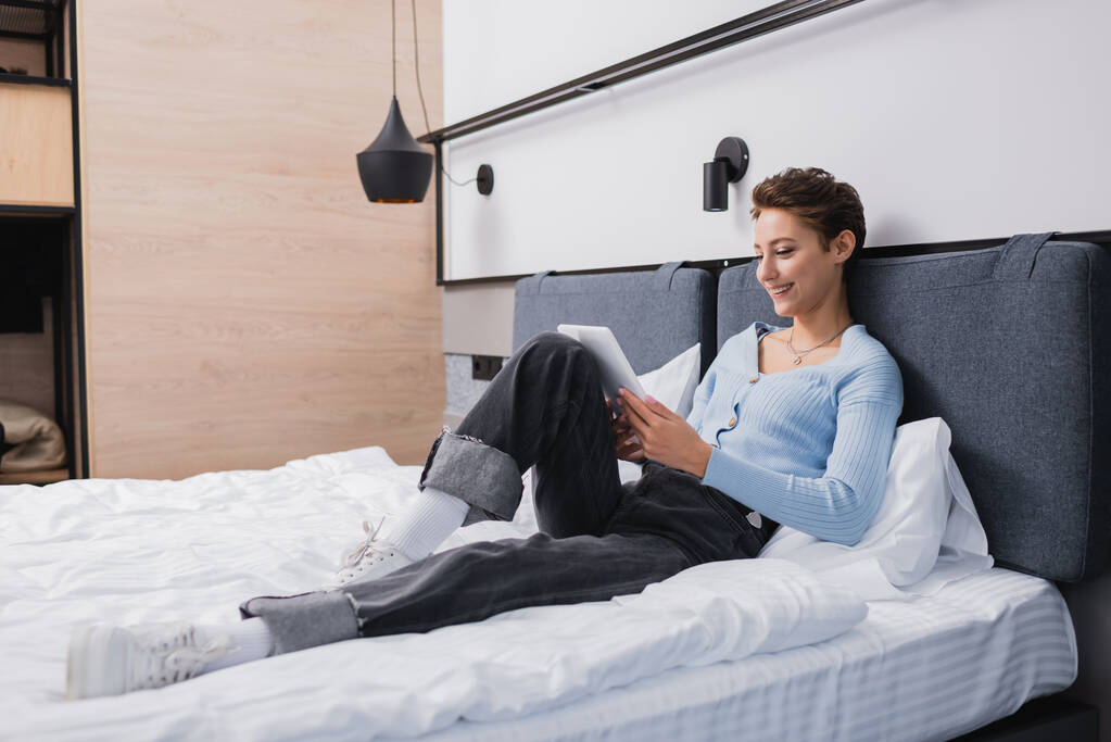 Otel odasındaki yatakta dijital tablet kullanan pozitif kadın.  - Fotoğraf, Görsel