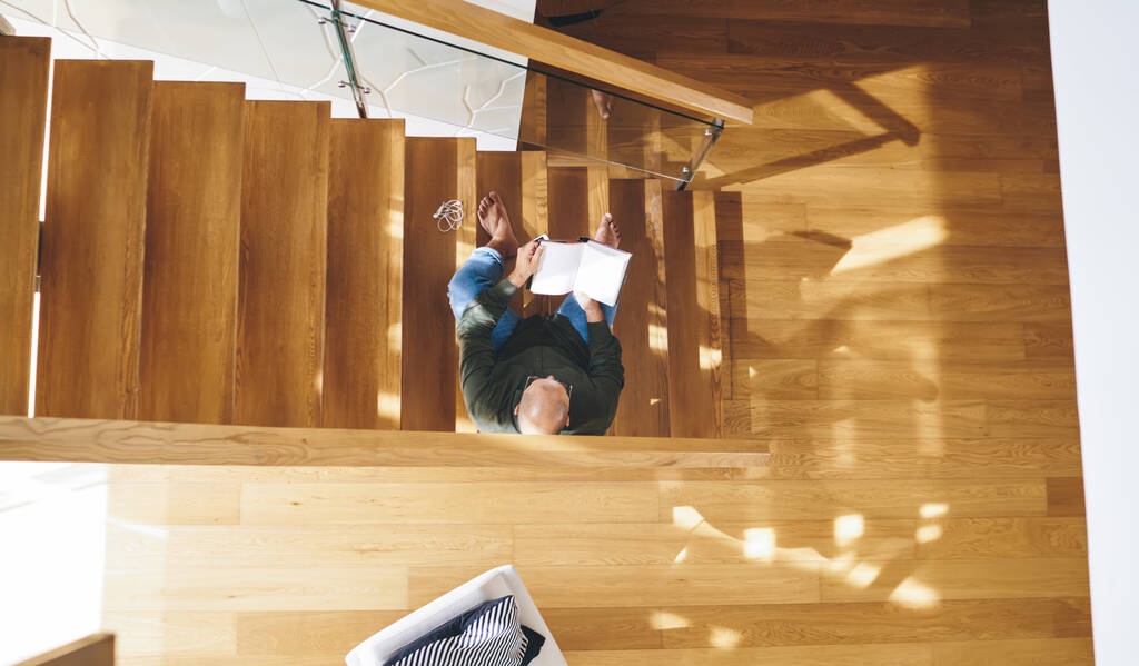 Vue du dessus du corps entier homme concentré en tenue décontractée assis sur l'escalier en bois et le livre de lecture tout en passant du temps dans un appartement moderne le week-end - Photo, image
