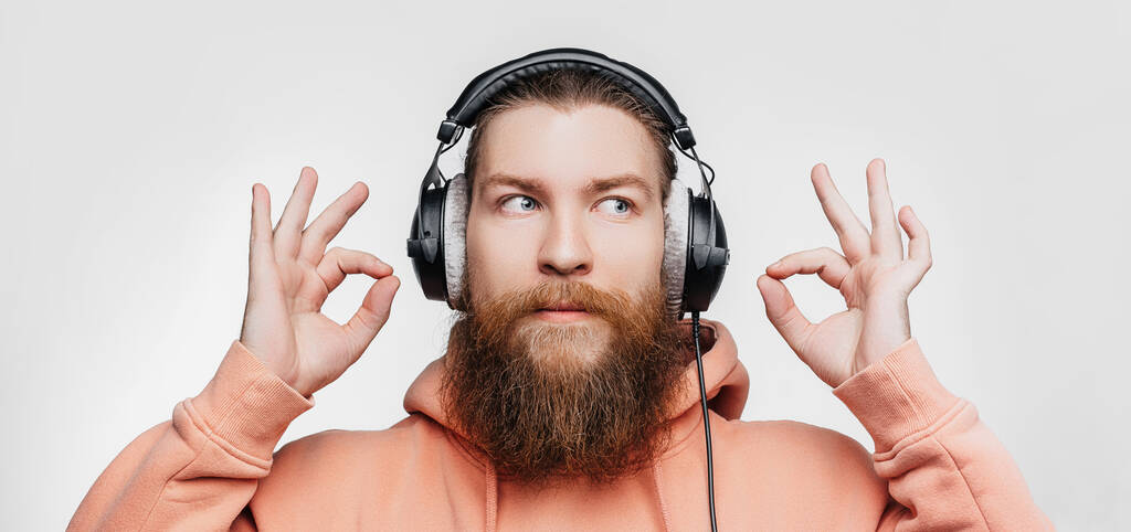 Scandinavische knappe man toont ok gebaar blijf kalm en luistert naar muziek in professionele koptelefoon geïsoleerd op grijze achtergrond. Gelukkige man met gember kapsel en baard. Moderne digitale technologieën - Foto, afbeelding