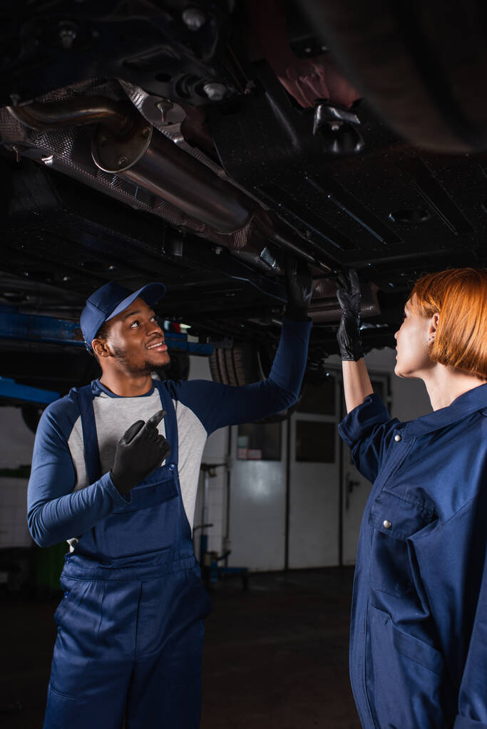 Smiling african american mechanic pointing at car bottom near colleague in service  - Foto, Imagem