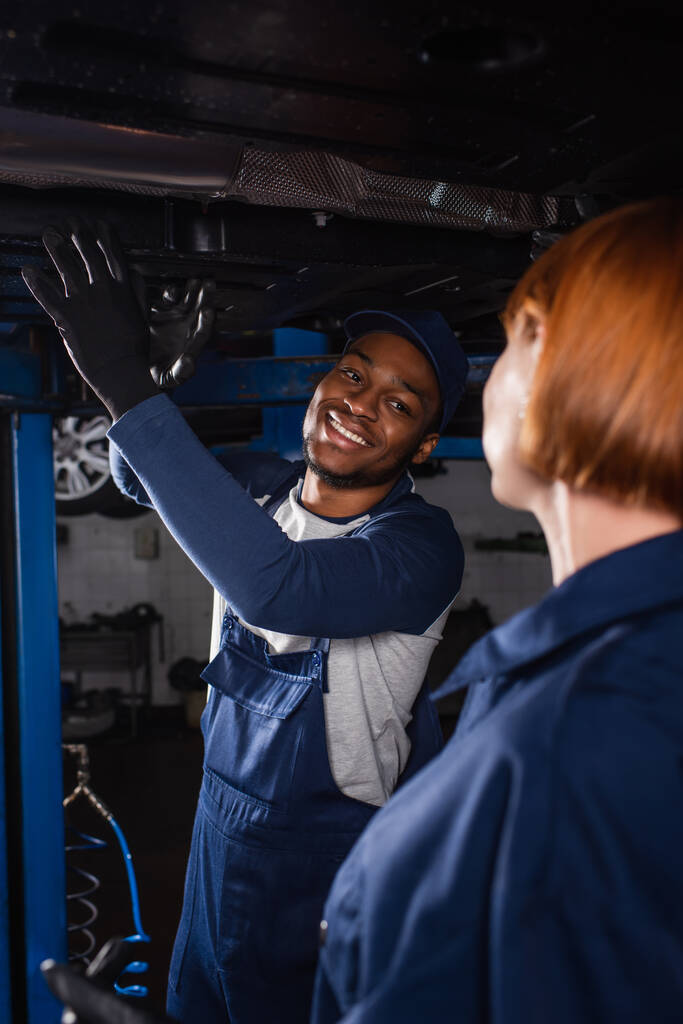 Sonriente afroamericano mecánico mirando borrosa colega cerca de auto en servicio  - Foto, imagen