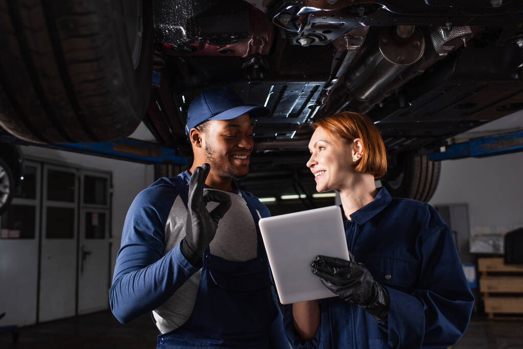 Χαμογελώντας αφροαμερικανός μηχανικός δείχνει ok χειρονομία κοντά στο συνάδελφο με ψηφιακή ταμπλέτα κάτω από το αυτοκίνητο στην υπηρεσία  - Φωτογραφία, εικόνα