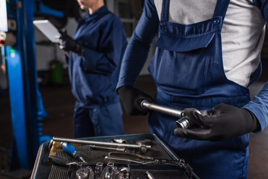 Ausgeschnittene Ansicht eines Mechanikers, der Schraubenschlüssel neben verschwommenem Kollegen mit digitalem Tablet in der Garage hält  - Foto, Bild