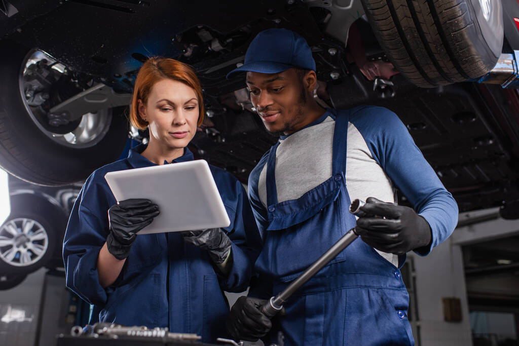 Multiethnic mechanics in gloves using digital tablet and holding wrench in car service  - Foto, Bild
