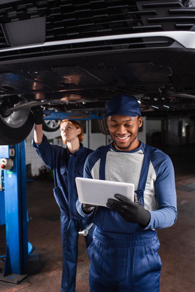 車のサービスでデジタルタブレットを使用して手袋でアフリカ系アメリカ人の機械工を微笑む  - 写真・画像