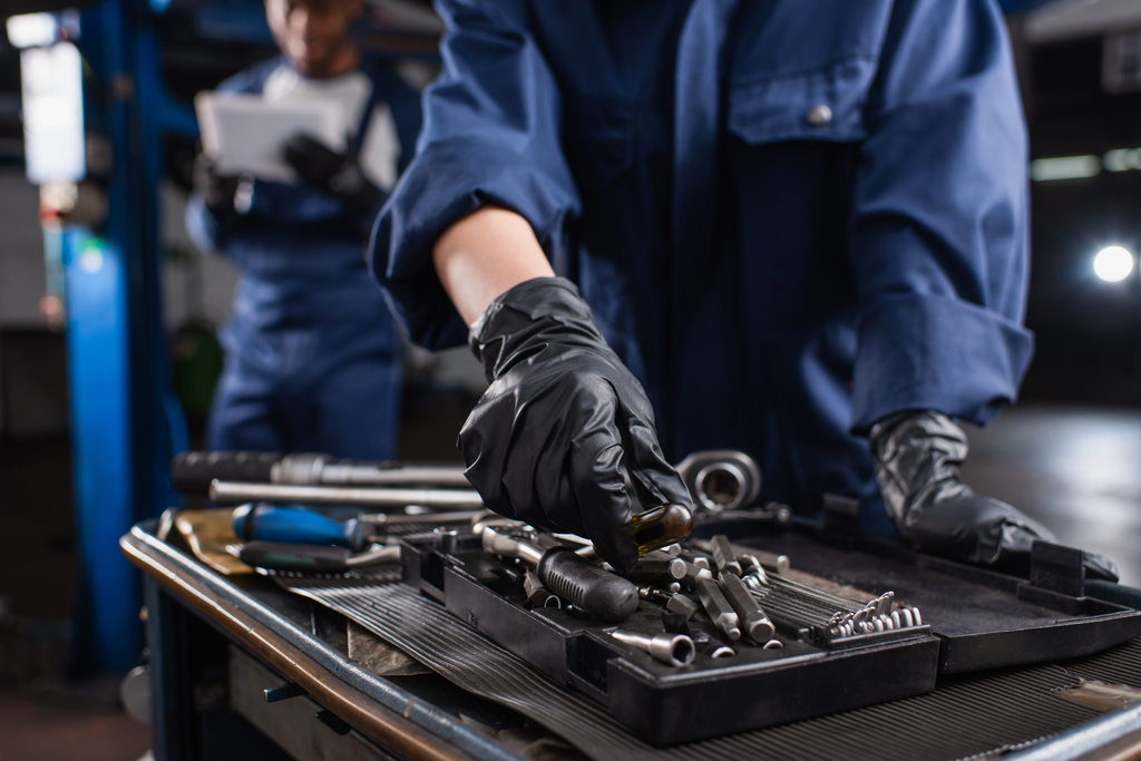 Przycięty widok mechanika w rękawiczkach podejmowania narzędzia w pobliżu zamazanego kolegi w serwisie samochodowym  - Zdjęcie, obraz