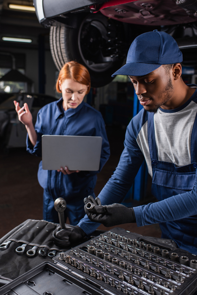 Африканский американский механик держит инструменты рядом с размытым коллегой, используя ноутбук в автомобильной службе  - Фото, изображение