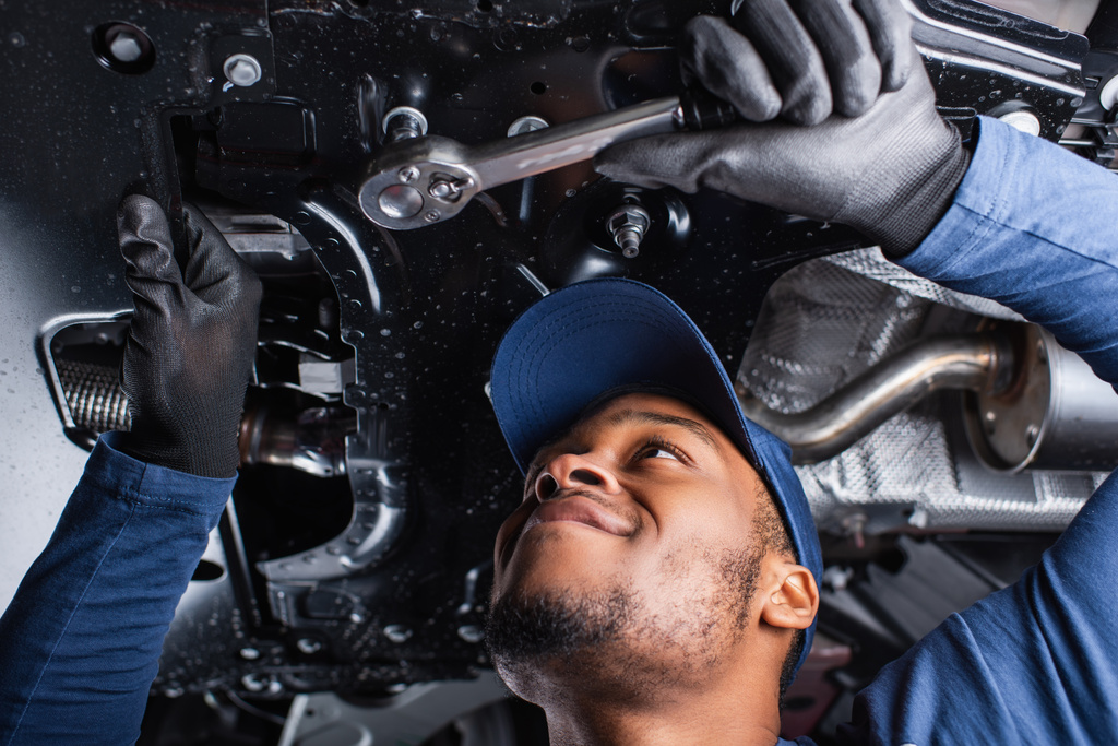 Nízký úhel pohledu veselý africký americký mechanik pracující s klíčem pod autem v garáži  - Fotografie, Obrázek