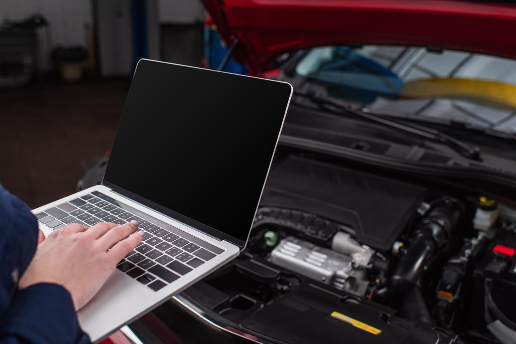 bijgesneden weergave van monteur met behulp van laptop met blanco scherm in de buurt wazig auto in de werkplaats - Foto, afbeelding