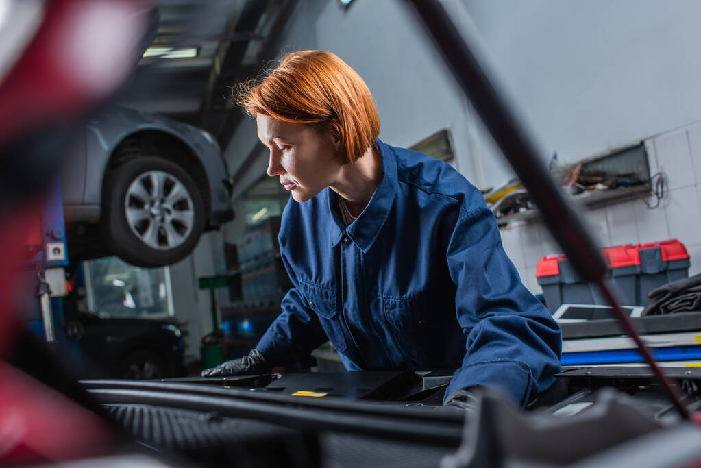 forewoman in uniform inspecting blurred car with open hood in workshop - Foto, Bild