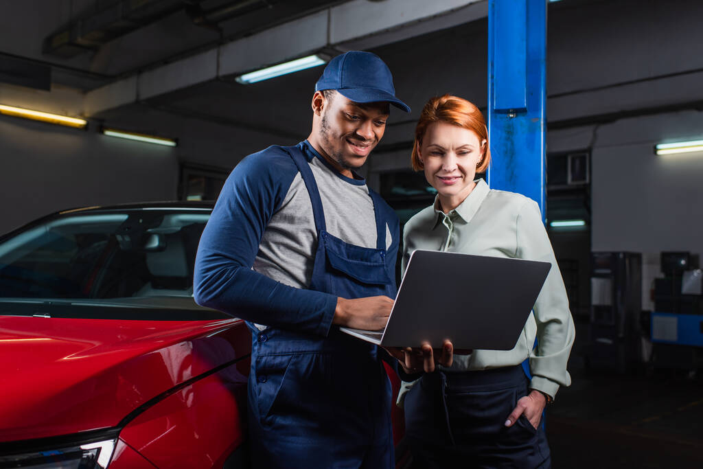 positieve Afro-Amerikaanse monteur en klant op zoek naar laptop in de buurt van auto in tankstation - Foto, afbeelding