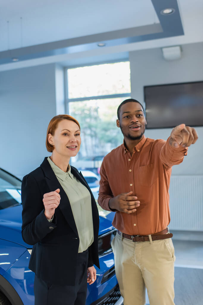 usmívající se africký Američan odvrací pohled a ukazuje prstem poblíž prodejce aut - Fotografie, Obrázek
