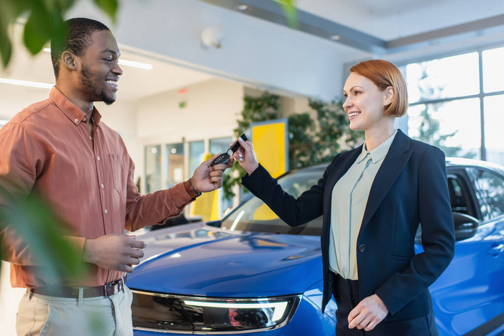 smiling car dealer giving key to joyful african american man in showroom, blurred foreground - Foto, Imagem