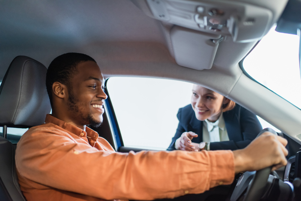 車に座っている陽気なアフリカ系アメリカ人男性の近くに手で指しているぼやけた車のディーラー - 写真・画像