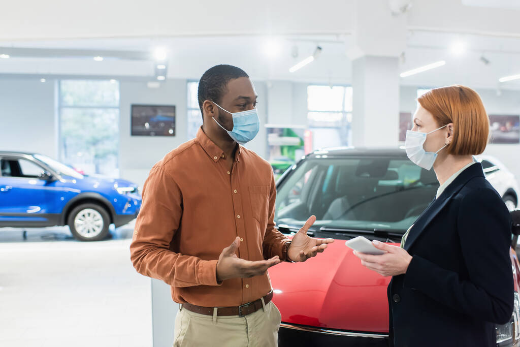 african american man in medical mask talking to manager of car showroom - Foto, Bild