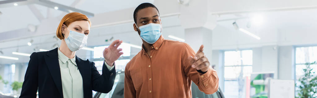 Afrikanisch-amerikanischer Mann und Autohändler in medizinischen Masken, die mit den Fingern im Autohaus zeigen, Banner - Foto, Bild