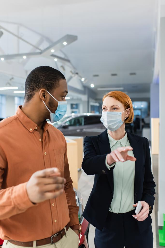 cliente afroamericano y concesionario de automóviles en máscaras médicas mirándose en la sala de exposición - Foto, Imagen