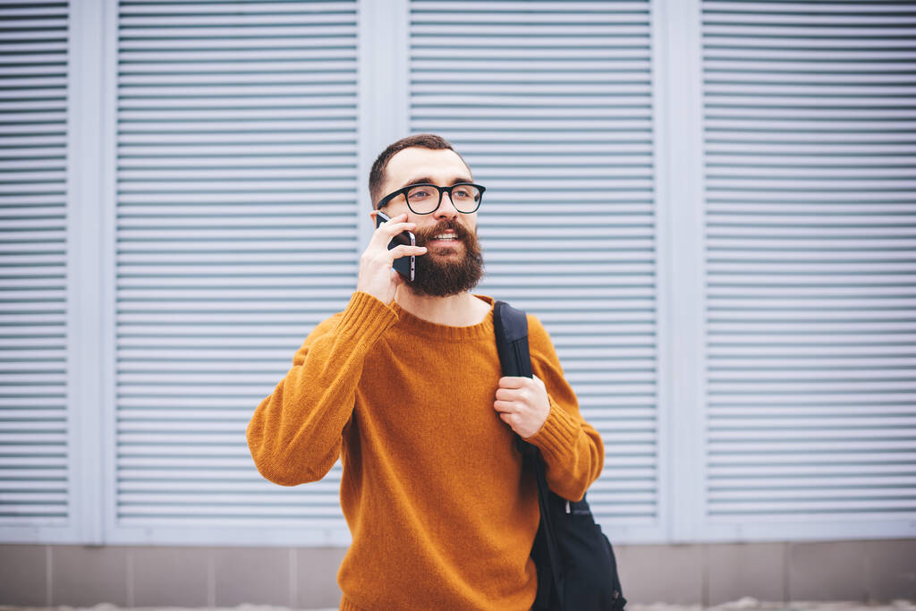 Όμορφος γενειοφόρος άντρας σε casual ντύσιμο με σακίδιο και γυαλιά κοιτάζοντας μακριά έχοντας συνομιλία στο smartphone έχοντας υπόλοιπο στο δρόμο - Φωτογραφία, εικόνα