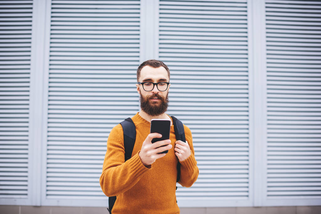Knappe hipster met baard in trendy brillenglazen en met rugzaknummer op mobiele telefoon naast gebouw met rolgordijnen op ramen - Foto, afbeelding