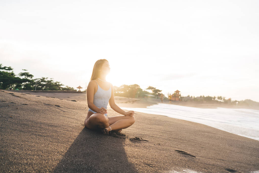 Volledige lengte van barefoot vrouwtje in badmode ontspannen op zandstrand in de buurt van rustig zeewater tijdens het mediteren tijdens heldere zonsondergang - Foto, afbeelding