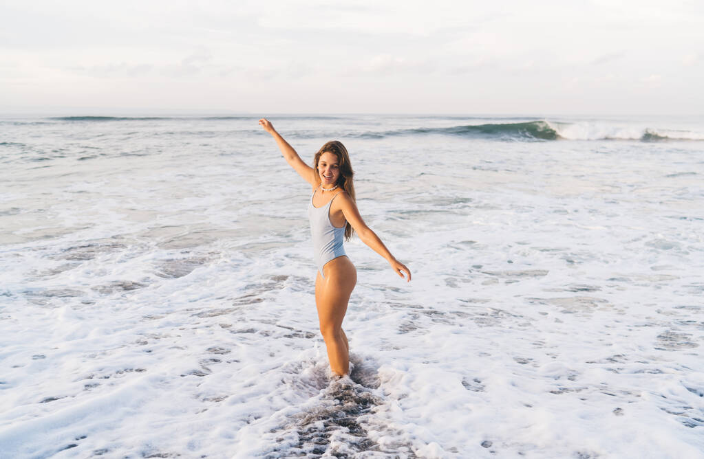 Вид збоку позитивна струнка жінка в білому купальнику, що стоїть з руками, витягнутими на морському узбережжі, вимитими пінистими морськими хвилями
 - Фото, зображення