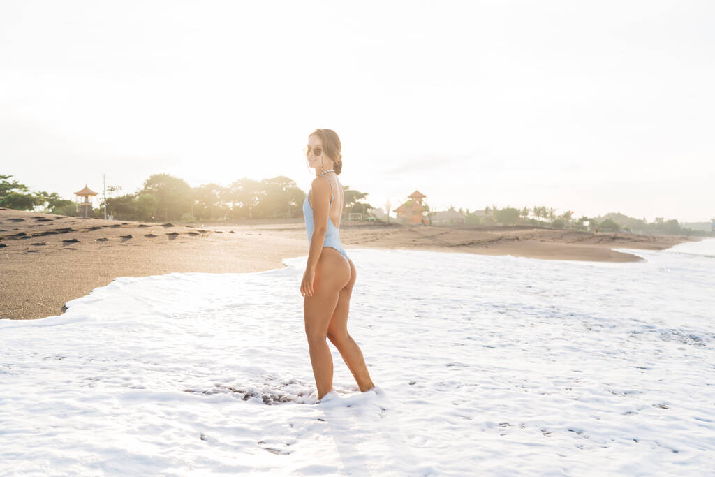 Вид збоку приваблива жінка з ідеальним тілом у стильному купальнику та сонцезахисних окулярах, що стоять на піщаному пляжі, вимитому пінистими морськими хвилями та дивлячись на камеру
 - Фото, зображення
