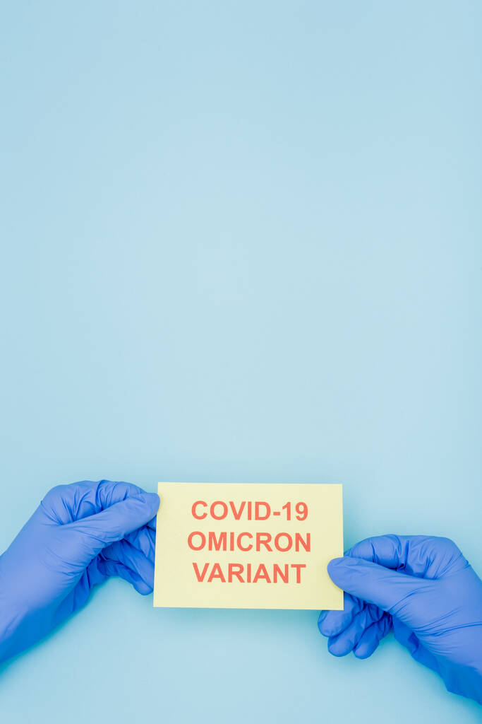 cropped view of doctor in latex gloves holding card with red covid-19 omicron variant lettering on blue - Fotografie, Obrázek