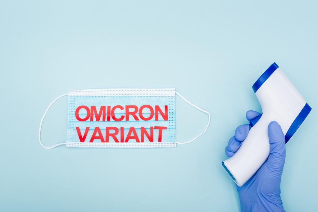 vue recadrée du médecin en gant de latex tenant pyromètre près du masque médical avec lettrage variante omicron sur bleu - Photo, image