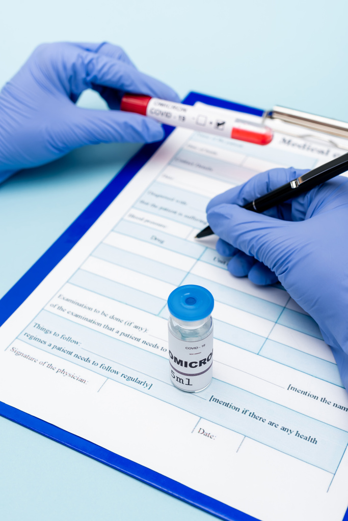 vue recadrée du médecin dans des gants en latex écrivant sur presse-papiers près du flacon avec covid-19 vaccin variante omicron sur bleu - Photo, image