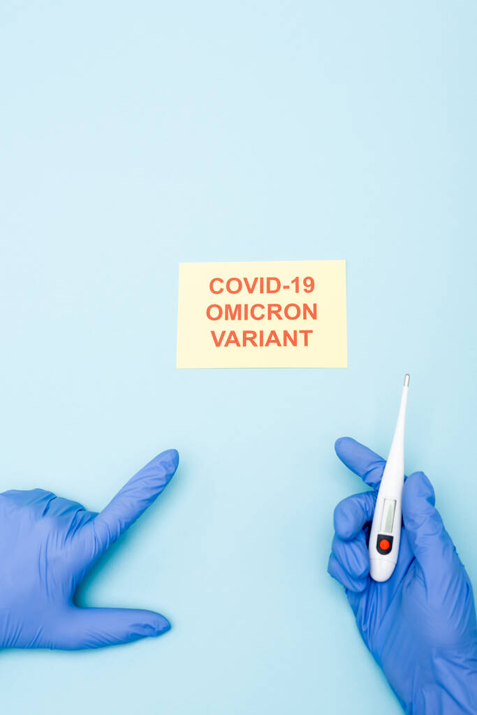 partial view of doctor with electronic thermometer pointing at card with covid-19 omicron variant lettering on blue - Foto, afbeelding