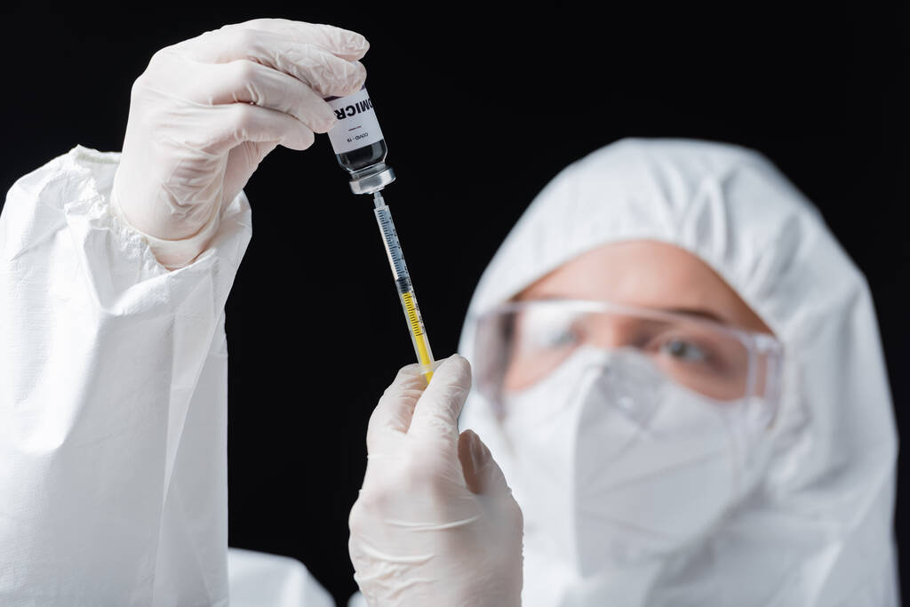 blurred immunologist in hazmat suit holding syringe and covid-19 omicron variant vaccine isolated on black - Photo, image
