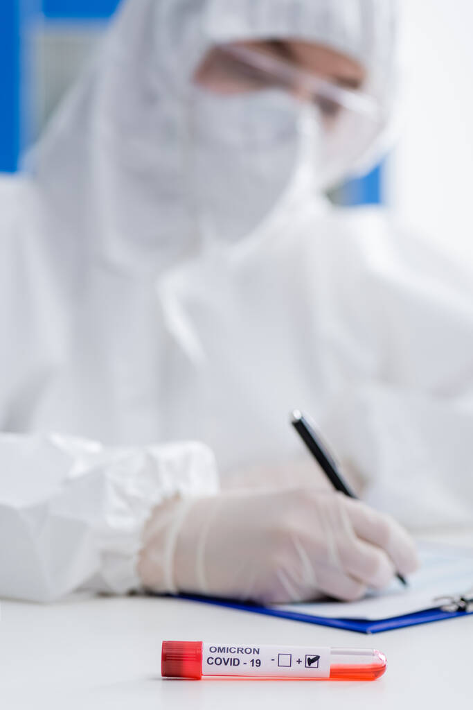 blurred scientist in hazmat suit writing on clipboard near positive covid-19 omicron variant test in lab - Fotografie, Obrázek