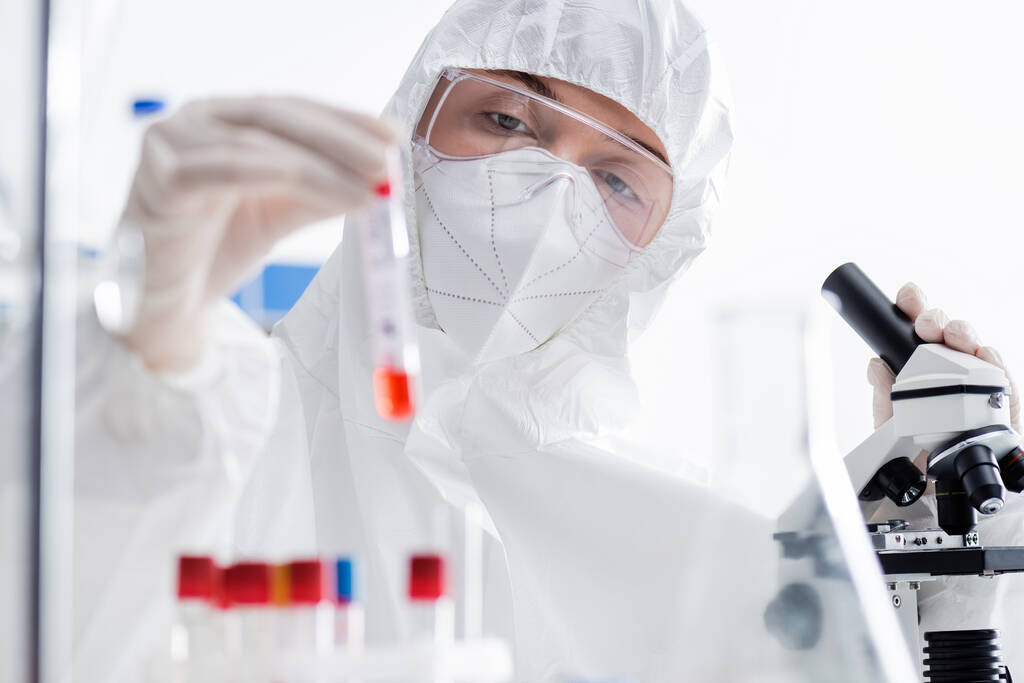 scientifique en lunettes, masque médical et combinaison Hazmat tenant un tube à essai flou près du microscope en laboratoire - Photo, image