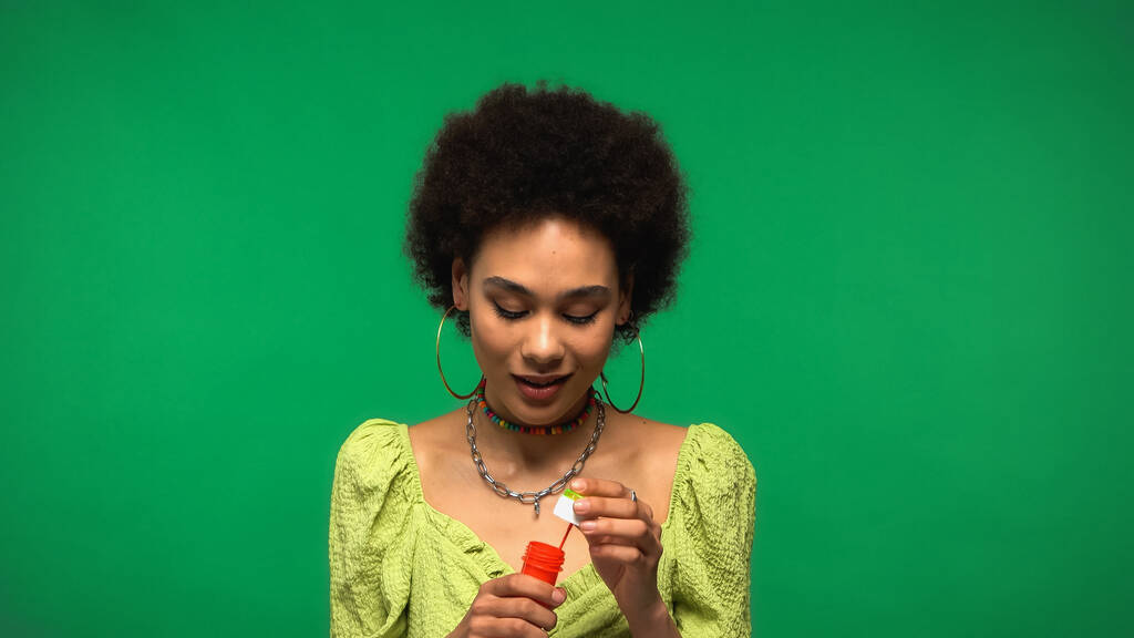 krullend Afrikaans amerikaanse vrouw houden fles met zeepbellen geïsoleerd op groen  - Foto, afbeelding