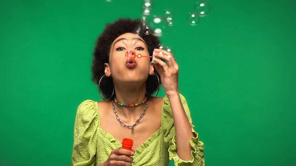 kudrnaté africké americká žena foukání mýdlové bubliny při držení bublina hůlka izolované na zelené  - Fotografie, Obrázek