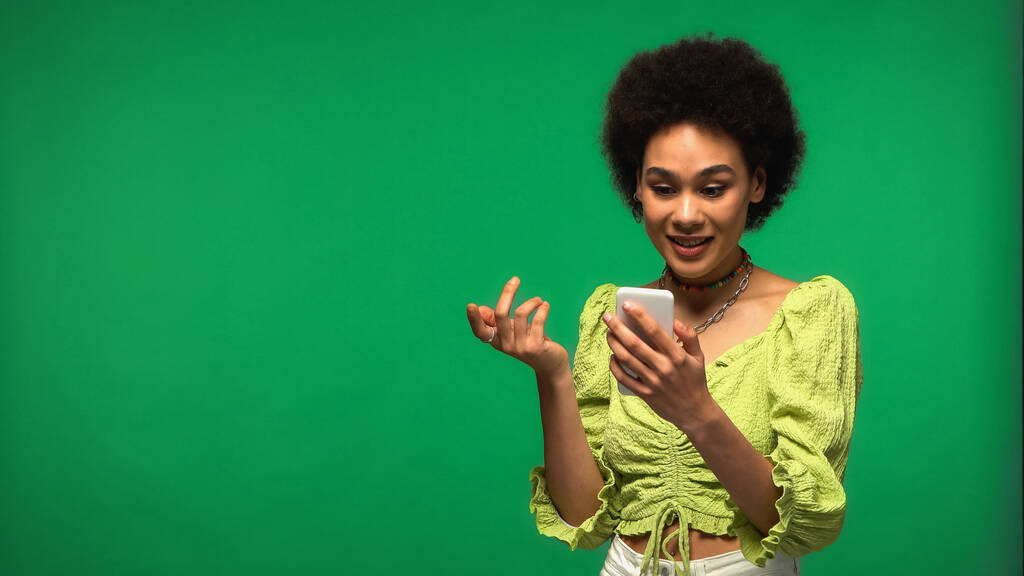 sonriente mujer afroamericana usando teléfono inteligente aislado en verde  - Foto, imagen
