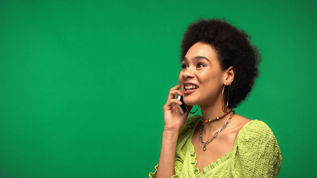 жизнерадостная африканская американка в блузке разговаривающая на смартфоне изолированная на зеленом  - Фото, изображение