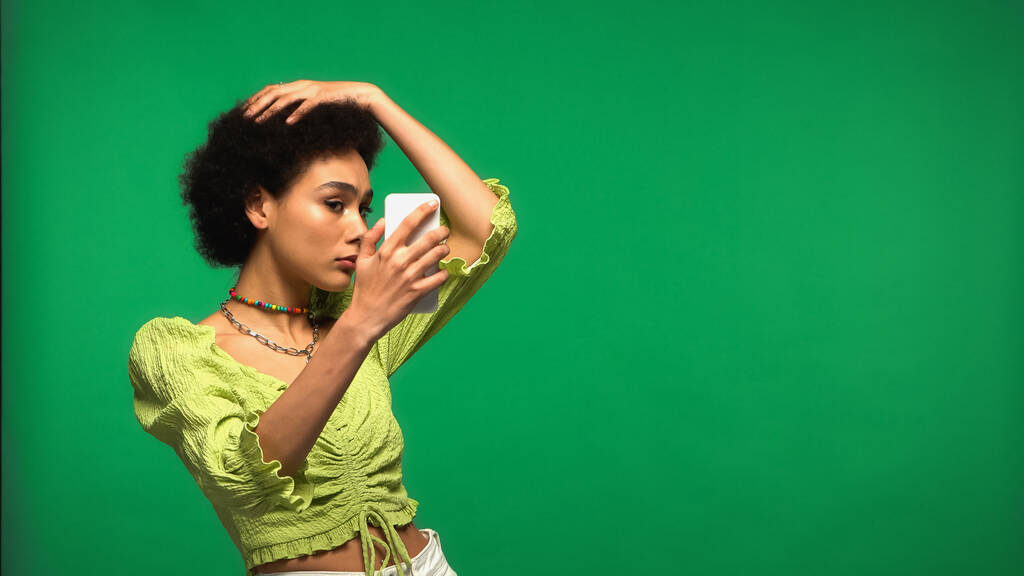 young african american woman in blouse looking at smartphone isolated on green  - Фото, зображення