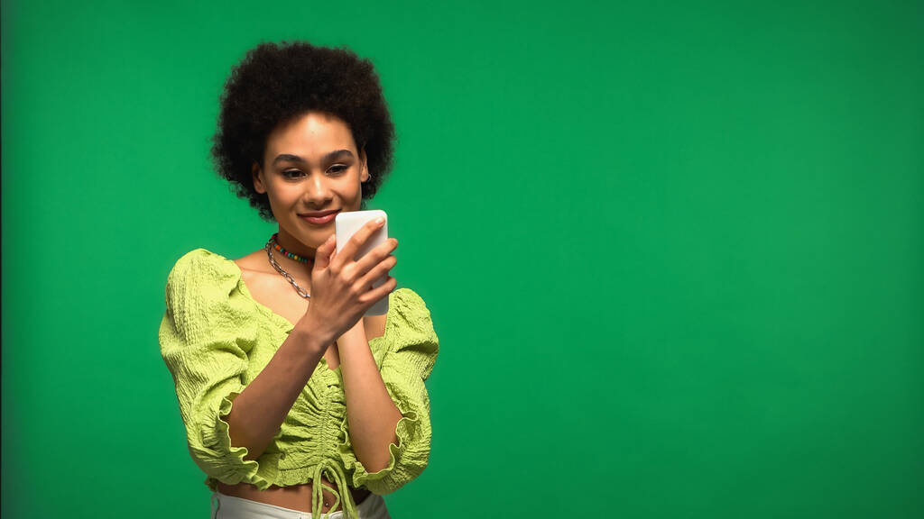 mujer afroamericana complacida en blusa mirando teléfono inteligente aislado en verde  - Foto, imagen