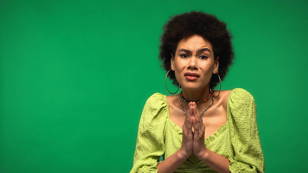 mujer afroamericana en pendientes de aro de pie con las manos de oración aisladas en verde - Foto, Imagen