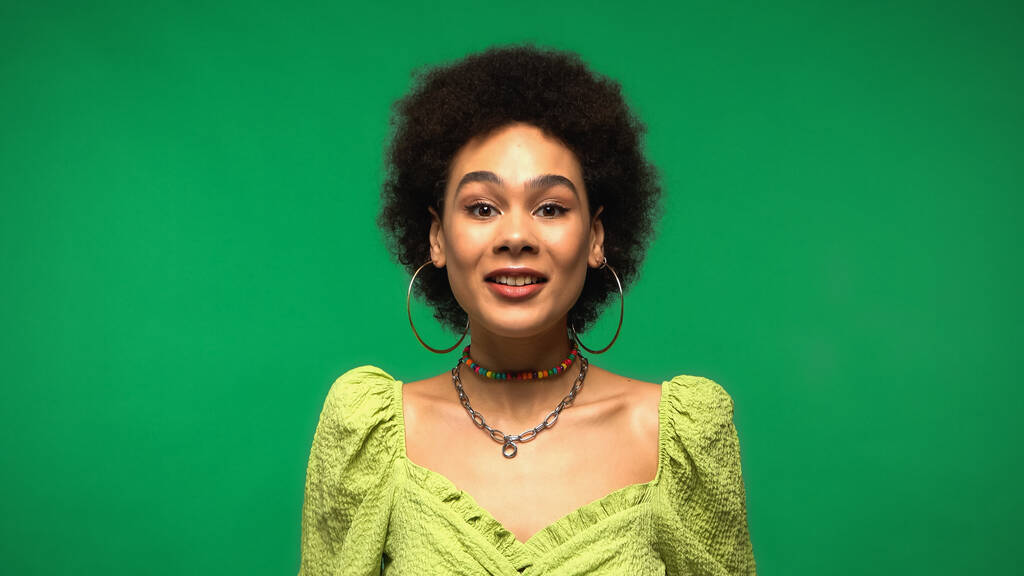 feliz joven afroamericana mujer en blusa y aros pendientes sonriendo aislado en verde  - Foto, Imagen