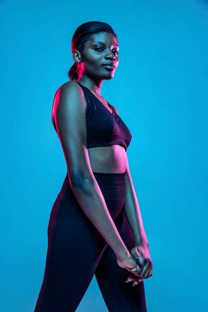Portrait of young beautiful dark skinned woman posing isolated on blue studio background in neon light. - 写真・画像