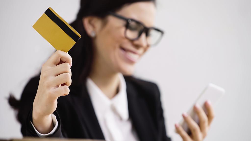 happy and blurred businesswoman doing online shopping while holding smartphone and credit card isolated on white  - Zdjęcie, obraz