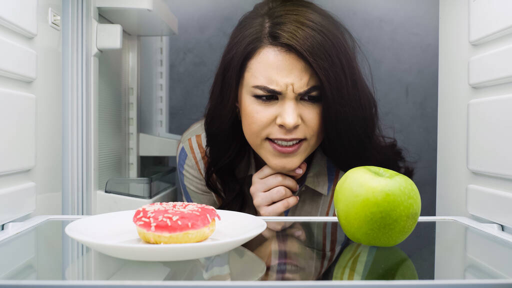 brunette woman choosing between apple and glazed doughnut in fridge  - Фото, изображение