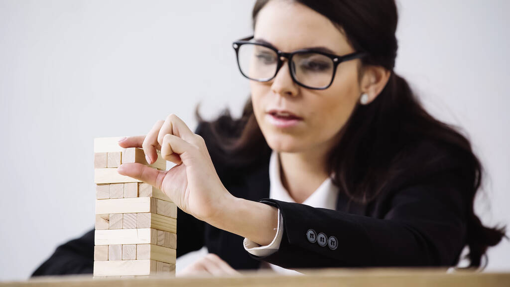 blurred businesswoman in glasses playing blocks wood tower game isolated on grey - Fotografie, Obrázek