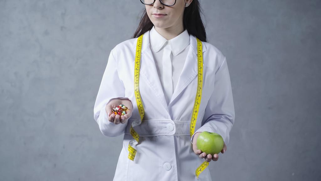 partial view of nutritionist in white coat comparing apple and vitamins on grey  - Foto, imagen
